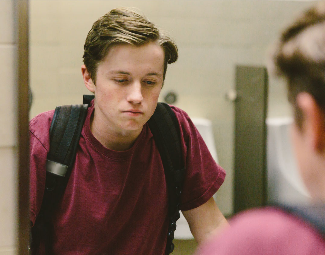 A teenage boy stands in front of a mirror in a school bathroom. His reflection is shown over his shoulder, and he looks defeated. 