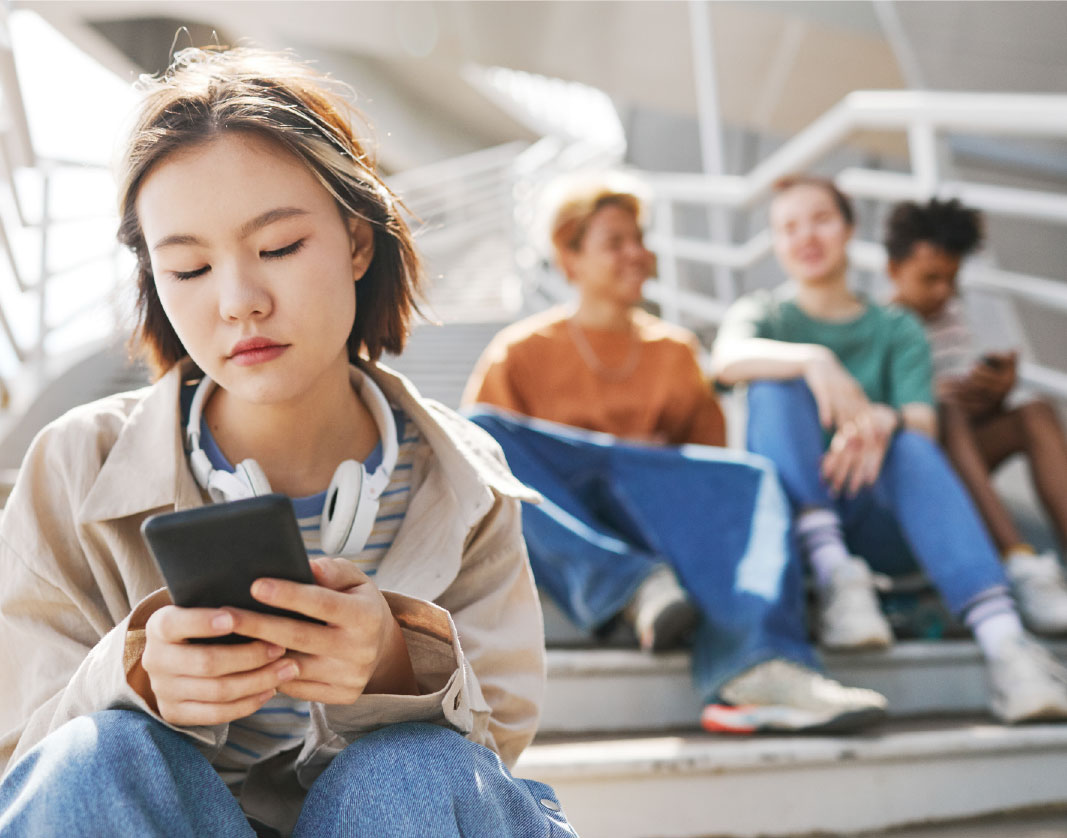 A teen girl sits on a staircase and looks at her phone and looks isolated. A group of teenagers sit behind her, out of focus.