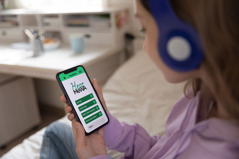 A young girl wearing headphones sits in her bedroom and looks at her phone, which has the HearMeWA app on screen.